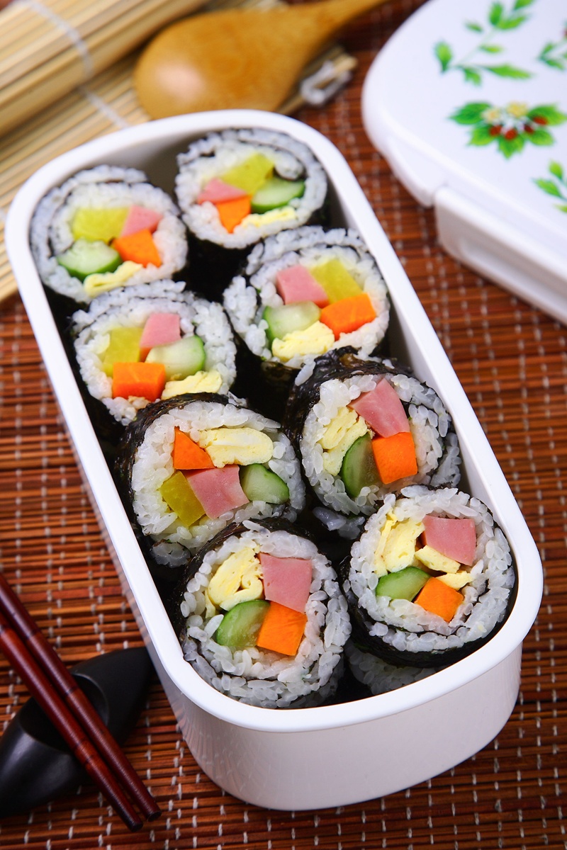 Sushi in einer weißen Box, gefüllt mit buntem Gemüse und Reisrollen auf Bambusmatte.
